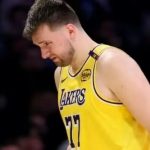 Lakers star Luka Dončić lowers his head in contemplation during a game break, clad in his iconic purple-and-gold No. 77 jersey, embodying the focus and self-reflection of a professional athlete amid intense competition.